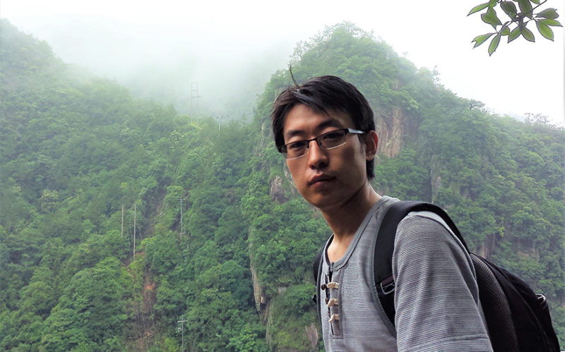 Bild på doktoranden Xin Luo framför ett högt berg med mycket växtlighet.